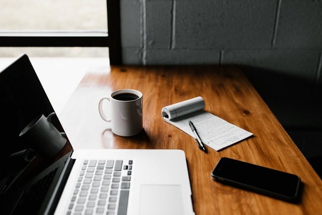 A laptop, notebook, phone and coffee sit on the desk ready to work