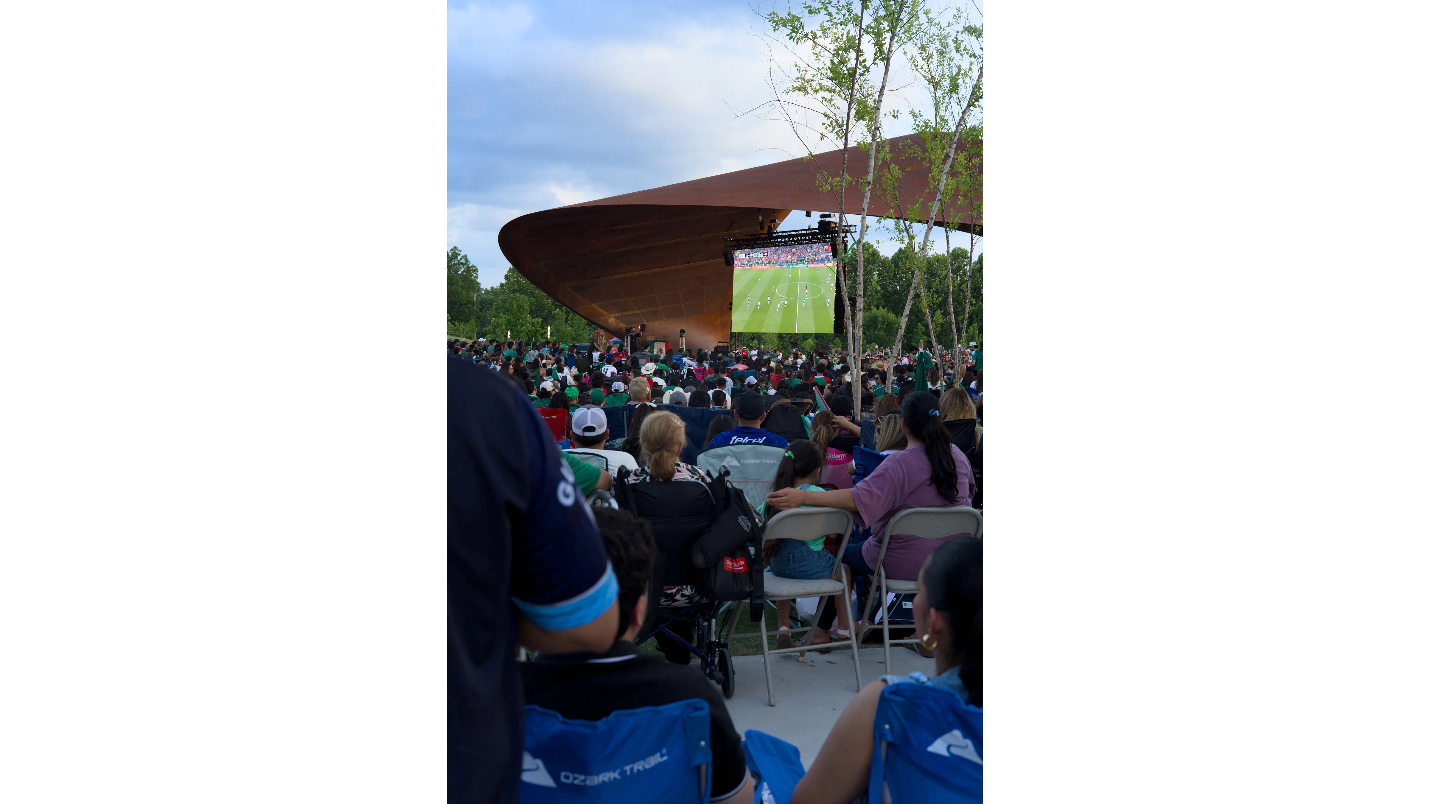 people sitting in park with trees on the side and pavilion with screen showing soccer