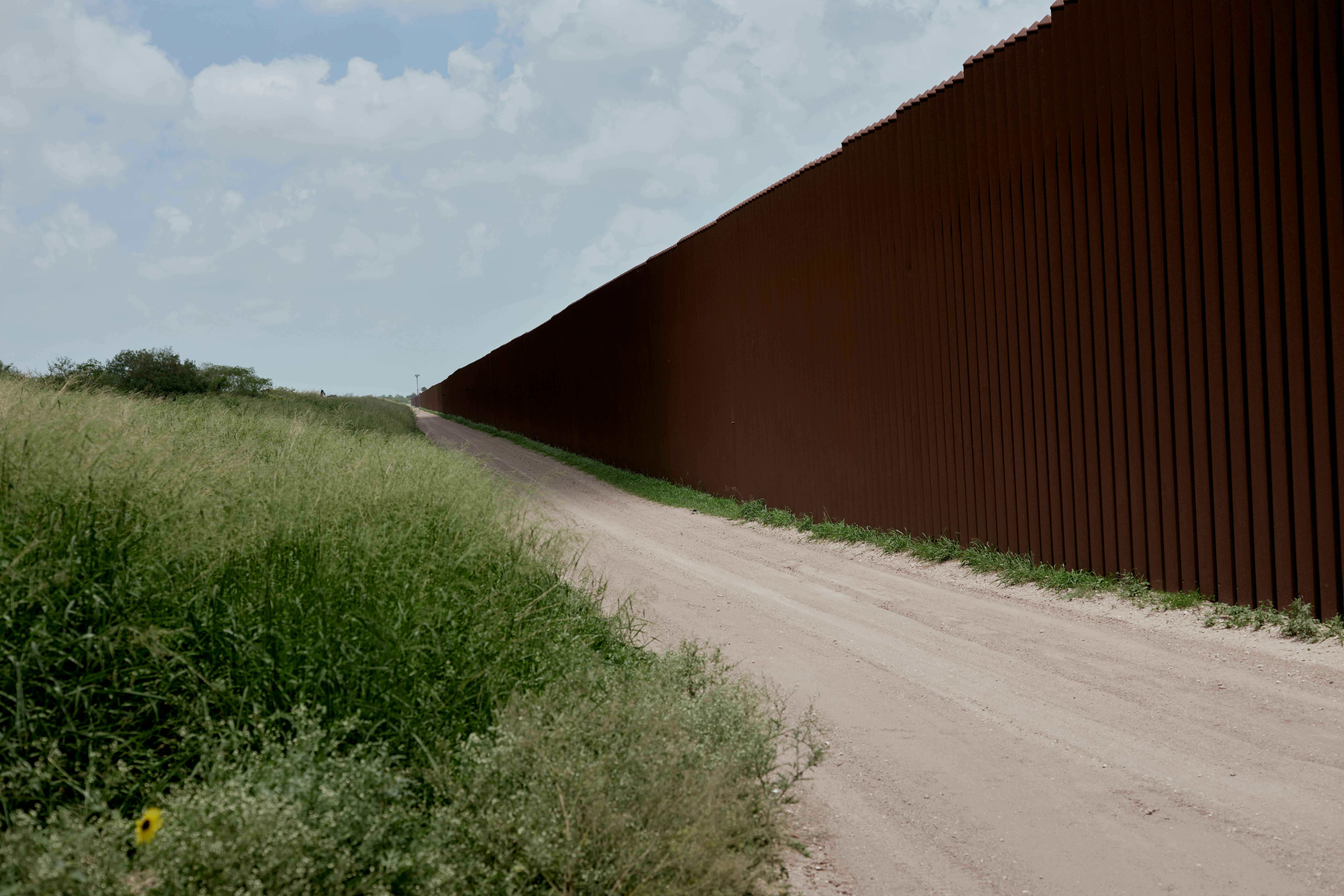US Mexico border wall along dirt road and field