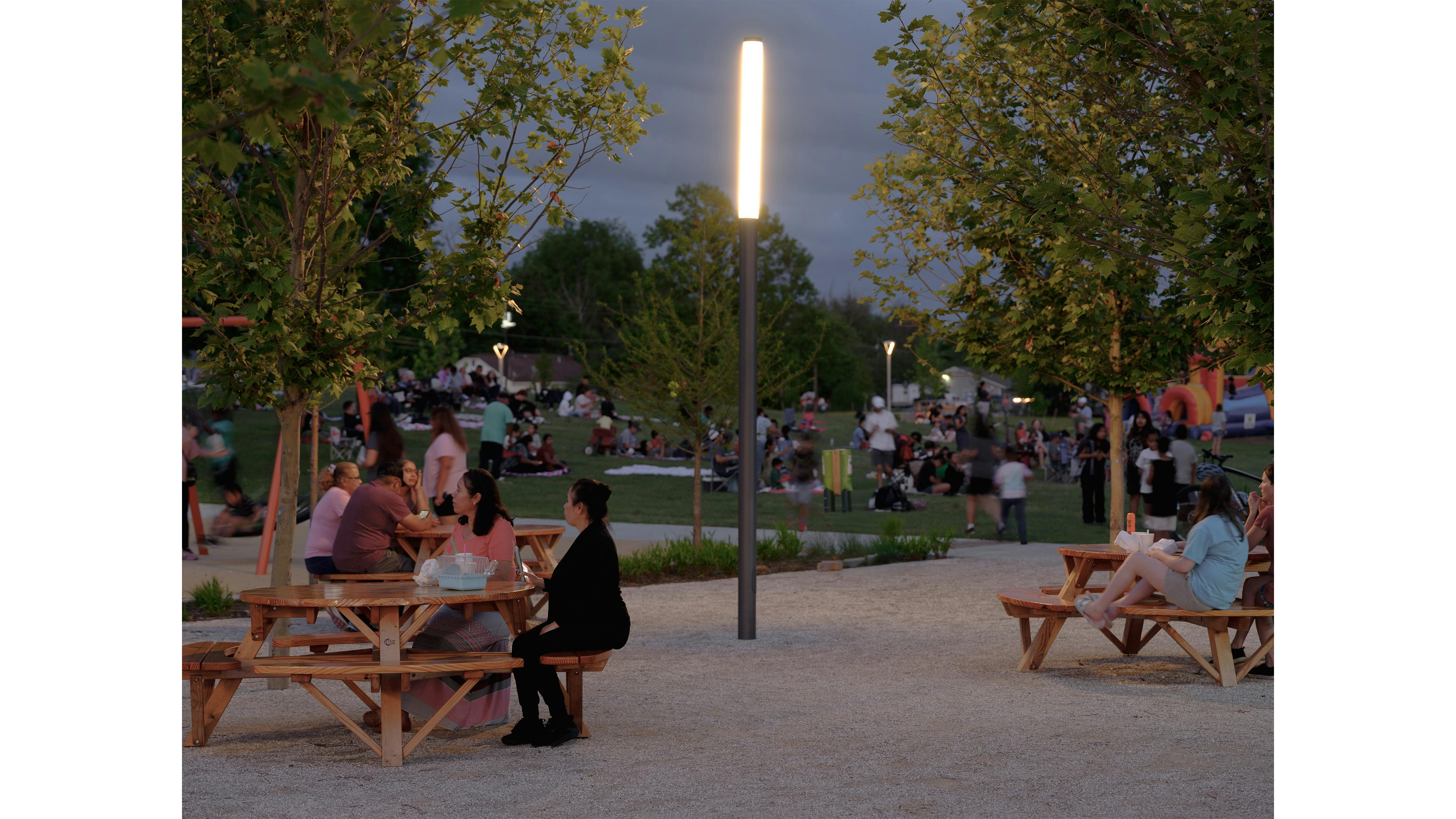families gather and sit at picnic tables in a park at dusk