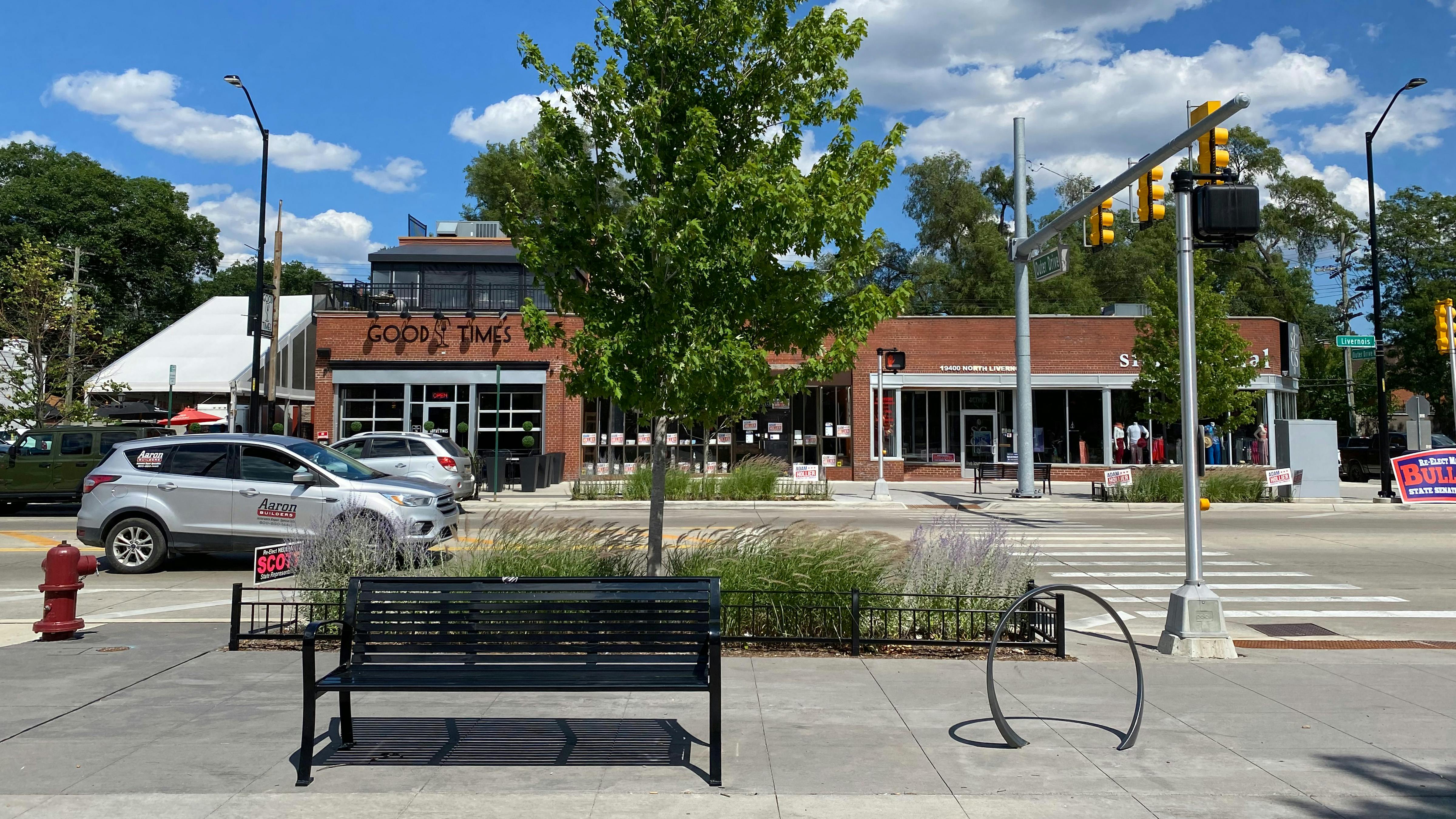Installed photograph of street bench and bike rack in front of bike lane, planting bed and new street trees dividing roadway in the background.