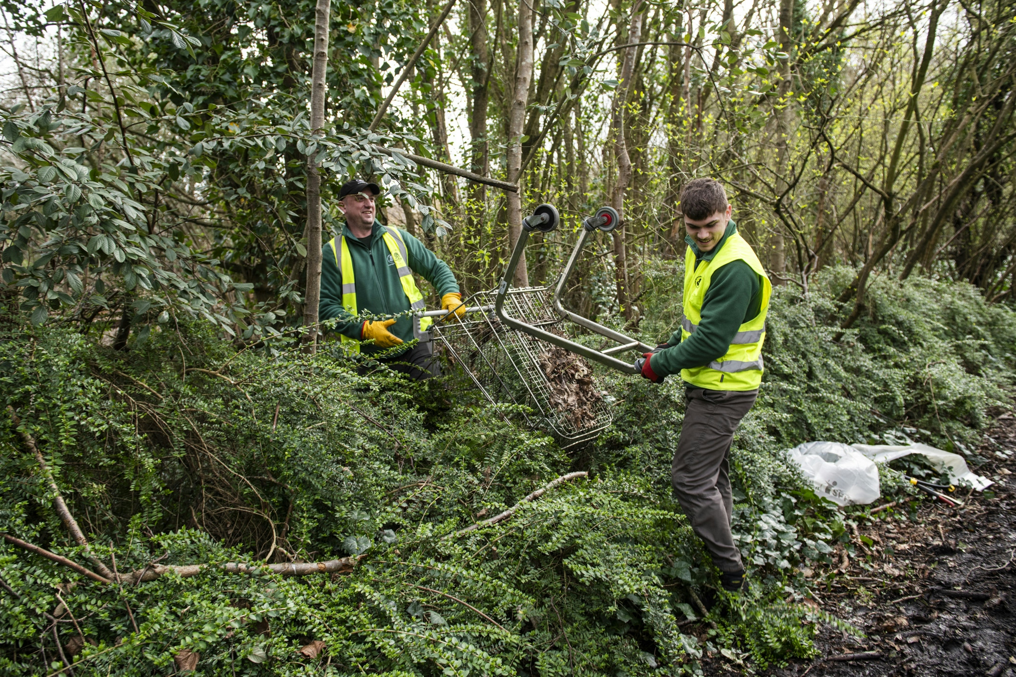 two men clearing a fly-tipped shopping trolley from a wooded area