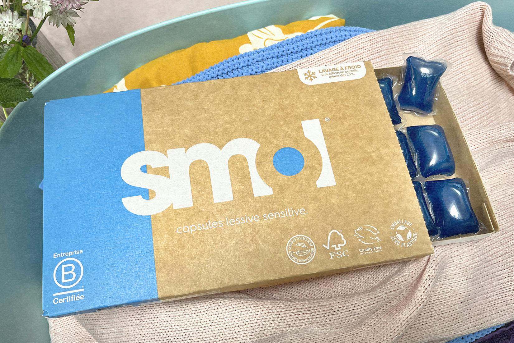 un pack de capsules de lessive sensitive dont le tiroir est ouvert, révélant quelques capsules de couleur bleue ; et posé sur une pile de linge de couleur dans un panier.