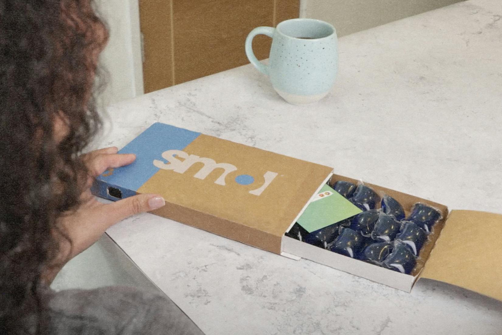 une femme aux cheveux bouclés chatains tenant un pack de capsules de lessive sensitive smol dont le tiroir est ouvert, révélant des capsules de couleurs bleue, sur une table en marbre à côté d’une tasse de café pleine.