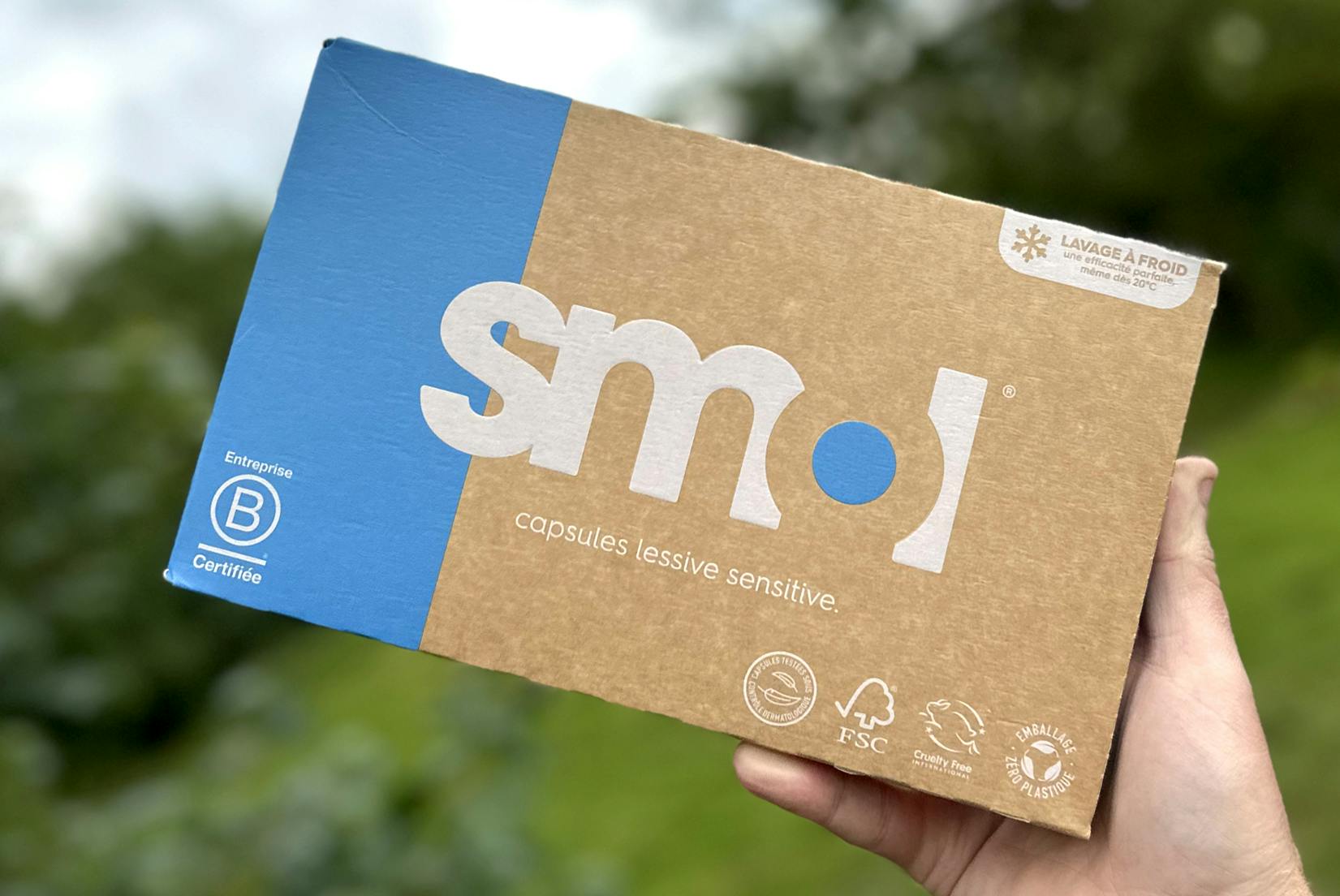 un pack de capsules de lessive sensitive smol à l’emballage marron et bleu avec des inscriptions et pictogrammes blancs, tenu en l’air par une main sur un fond de végétation.