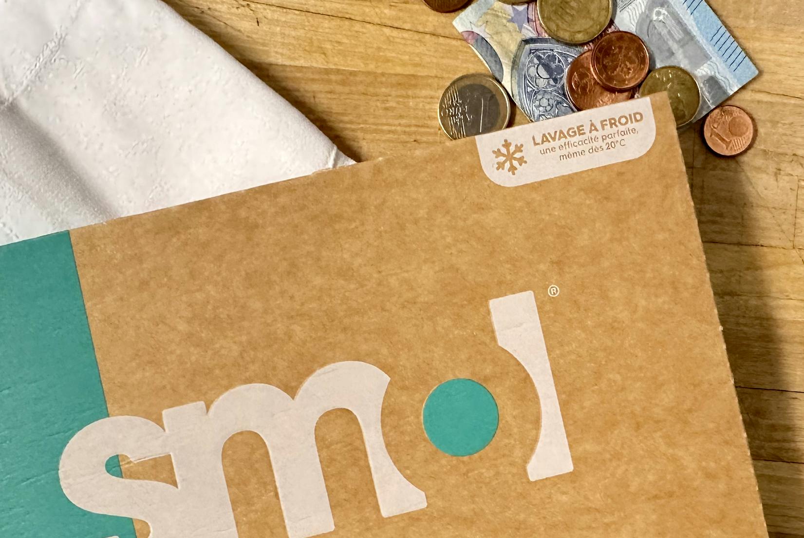 l’angle d’un pack de capsules de lessive active mentionnant « lavage à froid » et « une efficacité parfaite même dès 20 ºC » à coté d’un billet de 20 euros et de pièces de monnaie et d’un torchon blanc, le tout sur une table en bois.