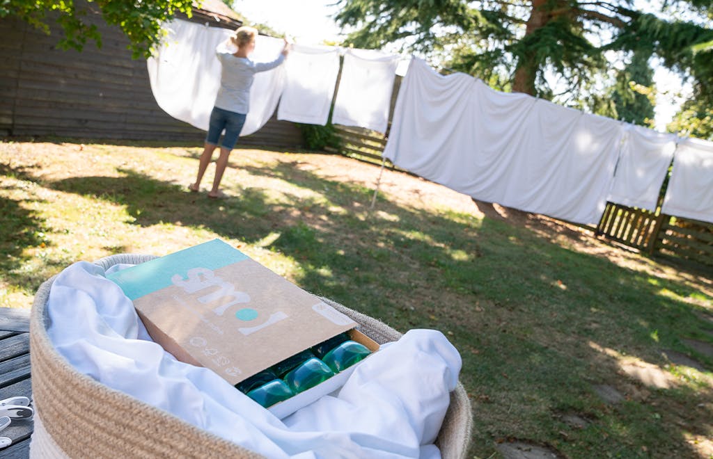 pack de capsules lessive active smol posé sur du linge blanc dans un panier de linge propre, dans un jardin avec un étendage et une femme qui étend le linge.