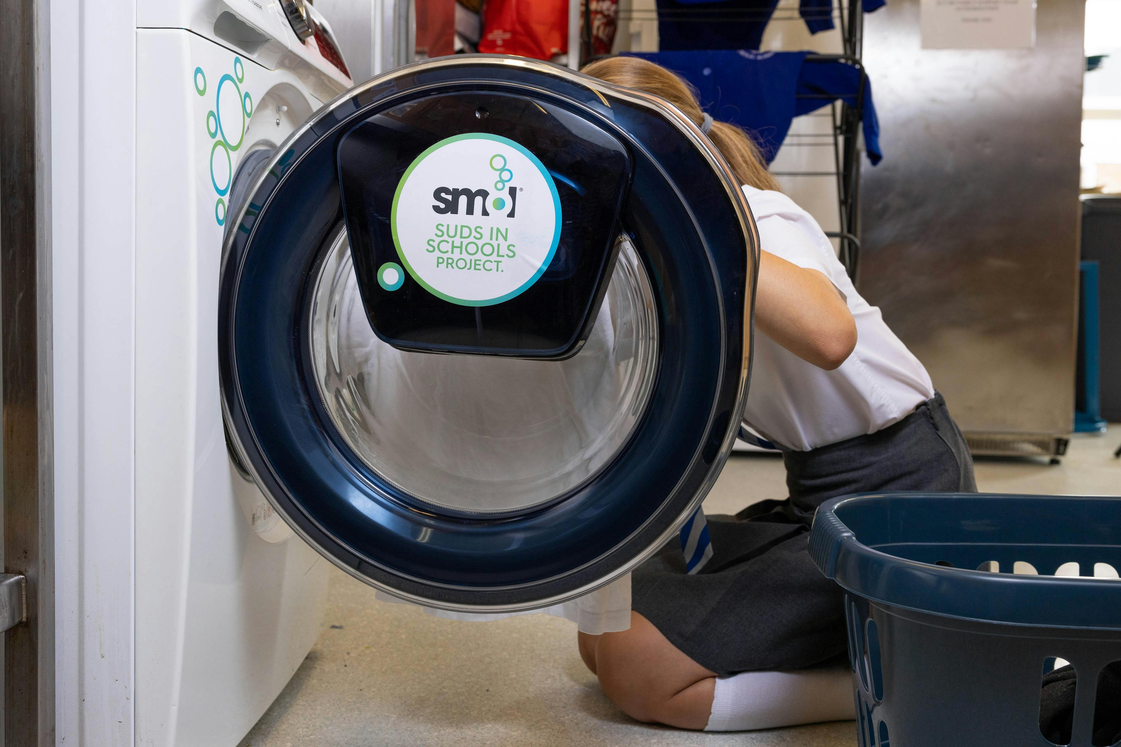 hublot d'une machine à laver ouverte avec une élève anglais remplissant la machine avec du linge. Le logo « smol suds in school » est sur le hublot.