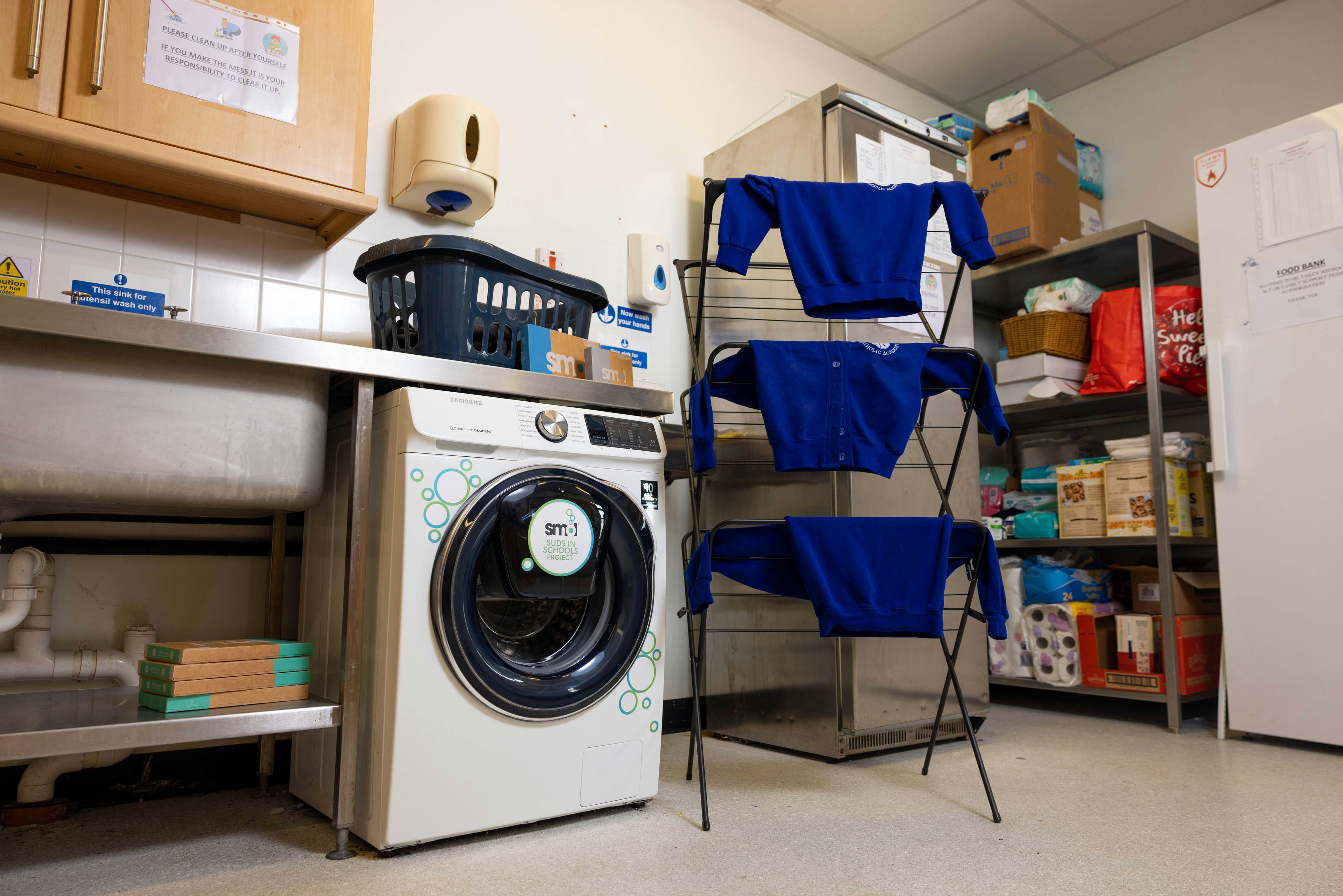 Buanderie Suds in School par smol dans une école britannique. Elle contient des étagères, une machine à laver et un étendage sur lequel sèchent des pulls d'uniformes d'élèves britanniques.