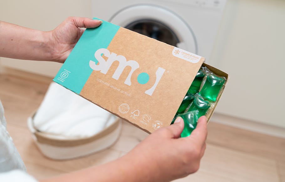 deux mains tenant un pack de capsules lessive active devant une machine à laver.