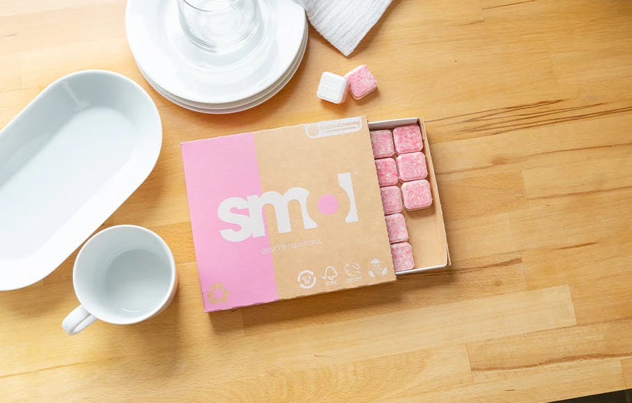 eine Packung smol Geschirrspültabs auf einem hellen Holutisch von oben fotografiert, daneben einige weiße Teller und Tassen