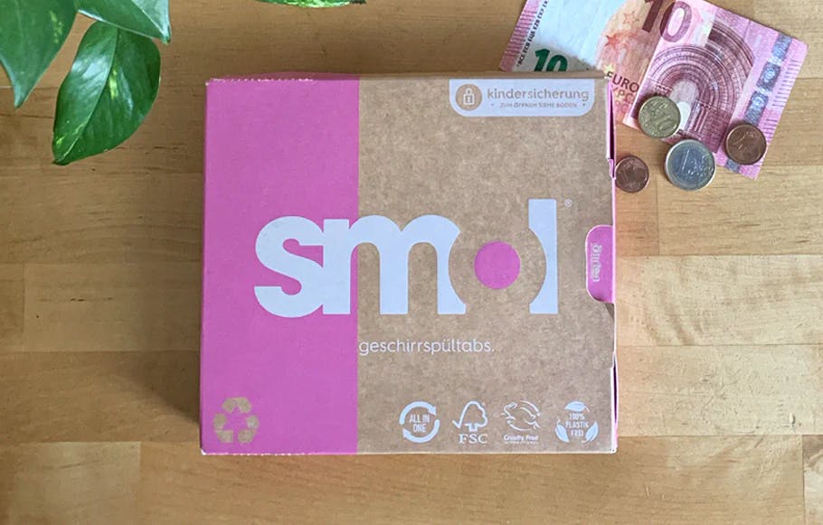 eine Packung smol Geschirrspültabs auf einem hellen Holztisch von oben fotografiert, daneben einige lose arrangierte Tabs und grüne Blätter