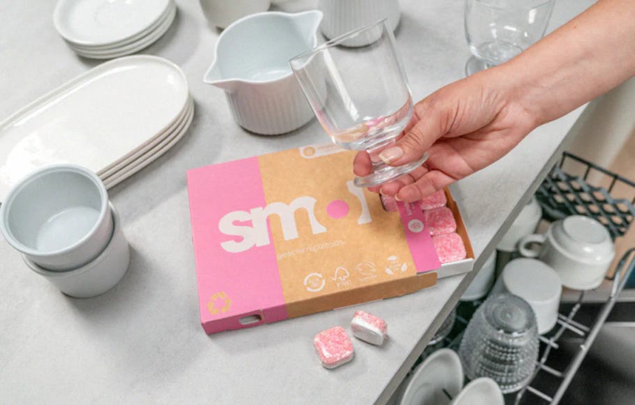 eine Packung smol Geschirrspültabs auf einer Küchenarbeitsplatte über einem geöffneten Geschirrspüler zwischen weißen Tellern und Tassen