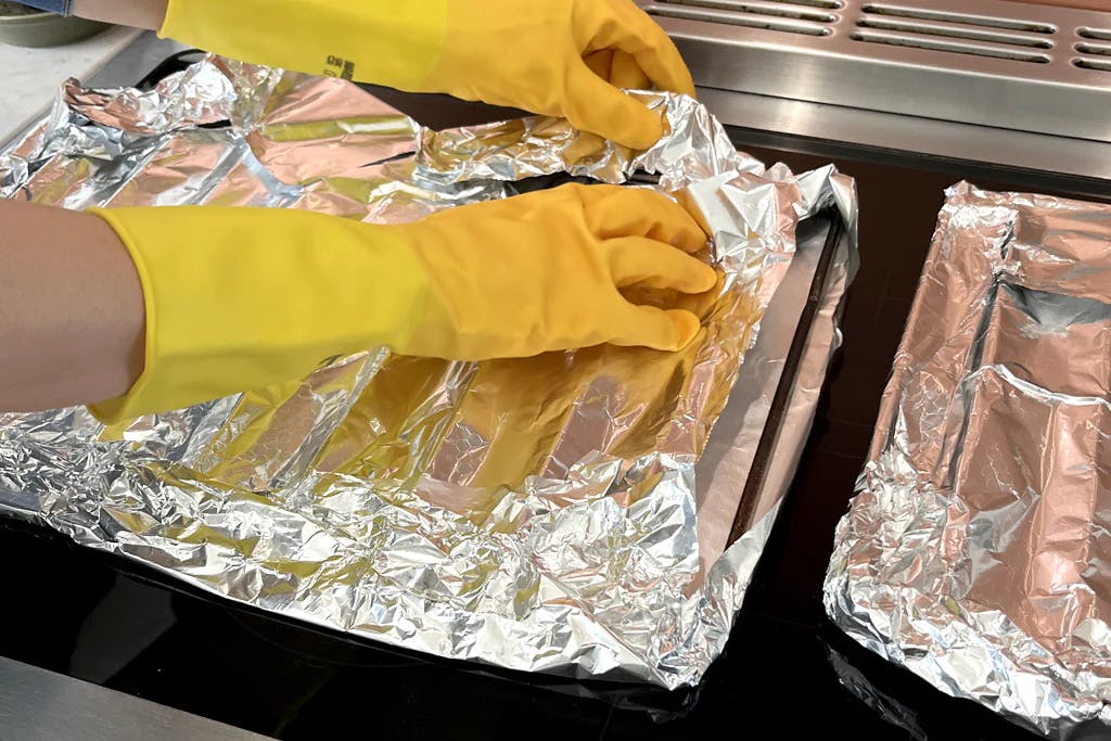 hands wearing rubber gloves wrapping aluminium foil around oven shelves