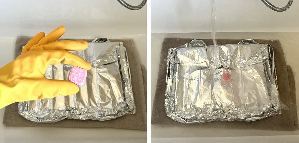 hand wearing rubber glove and holding a dishwasher tablet. Dishwasher tablet placed on top of oven shelves wrapped in foil, in a bath.