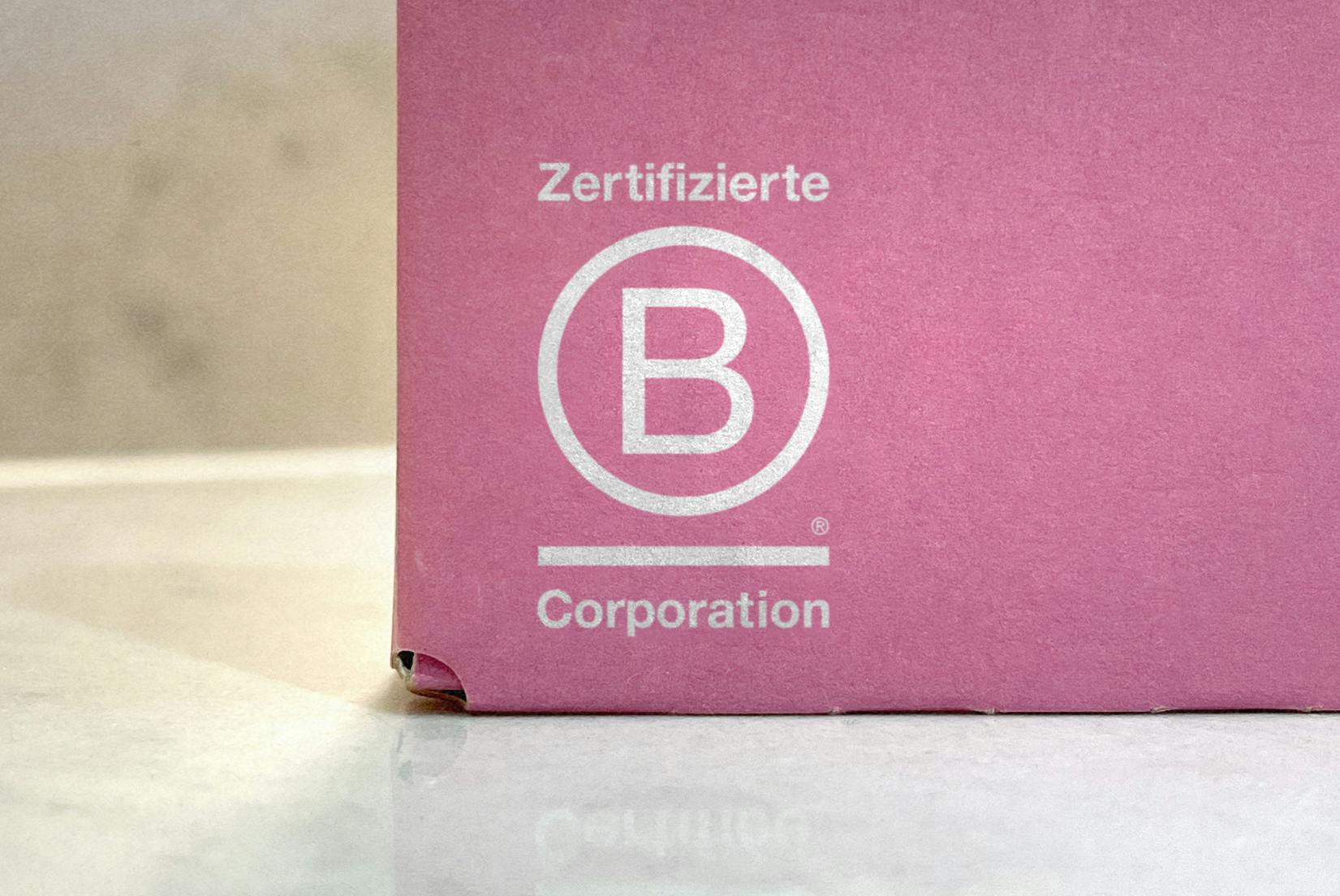 Das B Corp Logo in der utneren Ecke einer pinken smol Schachtel
