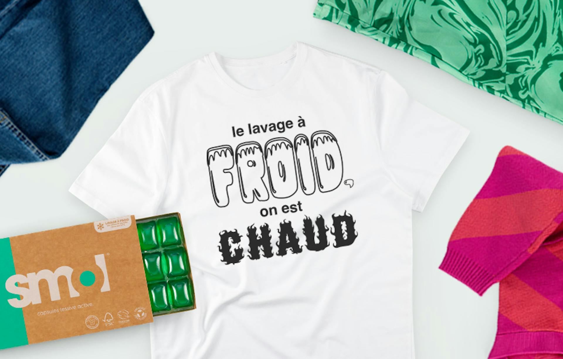 tee-shirt blanc avec le slogan « le lavage à froid, on est chaud » entouré d'un pull rayé rose et rouge, d'un tee-shirt vert et un tee-shirt bleu, posé à plat à côté de d'un pack de capsules lessive active avec le tiroir ouvert révélant des capsules vertes de lessive.