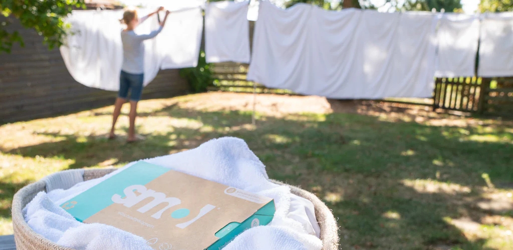 hanging laundry outside. laundry basket in the foreground filled with white towels and a box of smol laundry capsules on top.