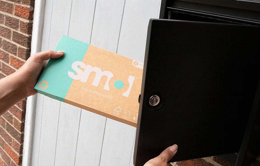 eine Packung smol Universal Kapseln, die aus einem schwarzen außen am Haus angebrachten Briefkasten entnommen wird