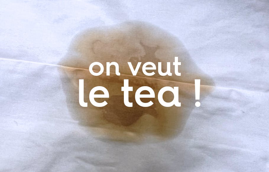 le texte « on veut le tea ! » sur une tache de thé sur un drap blanc.