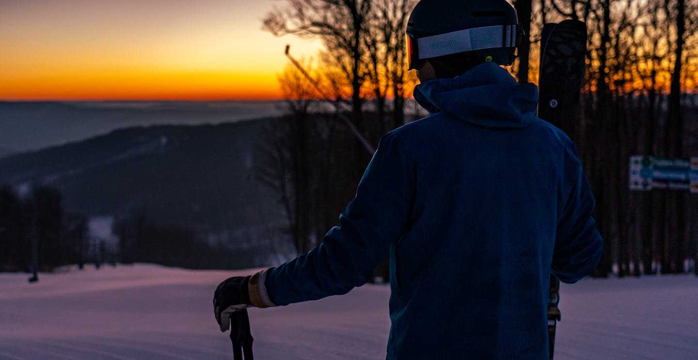 Male skier in blue ski jacket admiring the sunrise from the slopes at Holiday Valley in New York, USA.