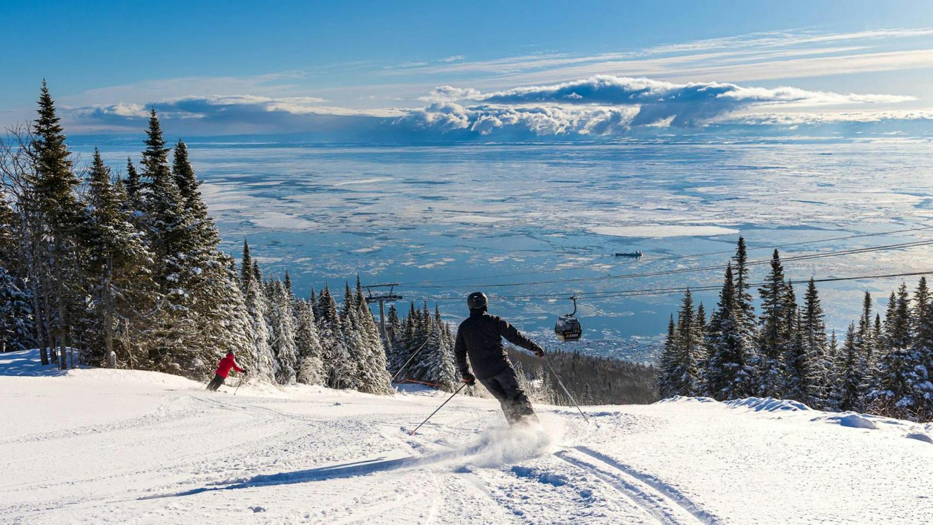 Two skiers of undiscernible gender hitting the slopes while the gondola runs over the St. Lawrence River at Le Massif de Charlevoix near Montreal in Quebec, Canada.