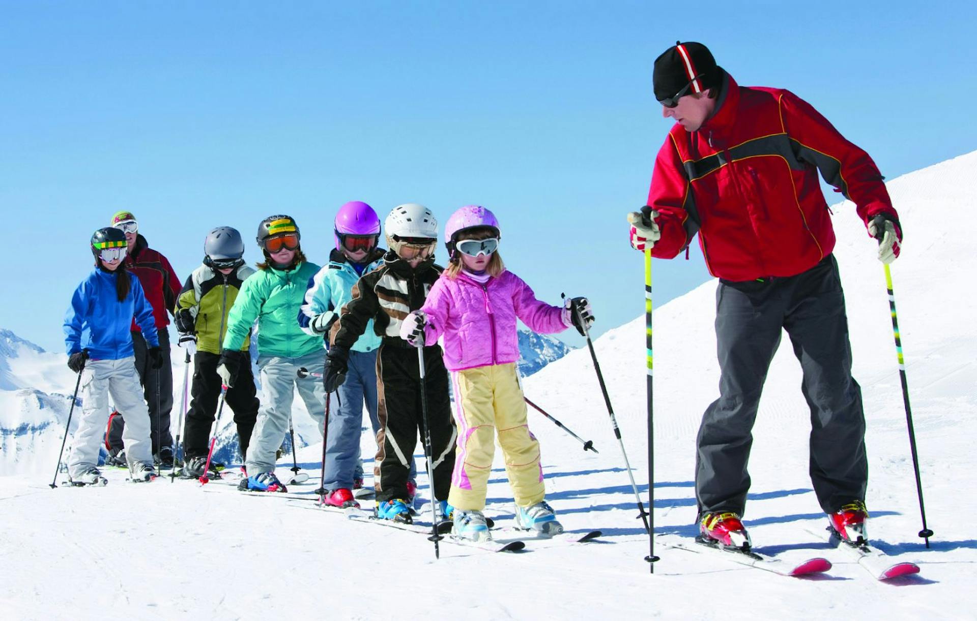 Two male Lookout Pass ski instructors in red ski jackets giving a ski lesson to a diverse group of six children in colorful ski attire at Lookout Pass in Idaho, USA.