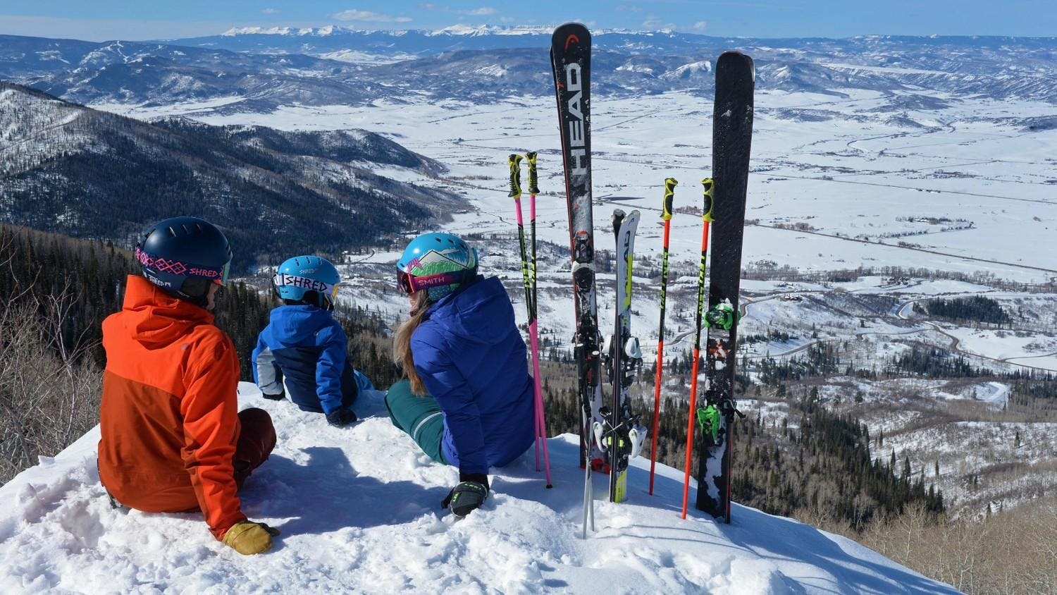 Three skiers in orange and blue ski attire resting on the slopes at Steamboat Springs in Colorado, USA.