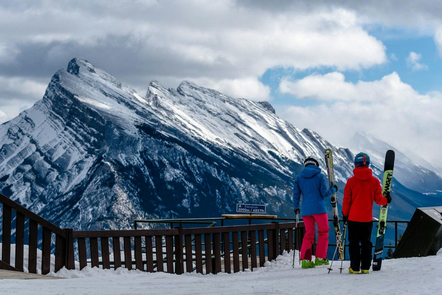 Two skiers of undiscernible gender in brightly-colored ski attire gazing at the peak at Mount Norquay in Alberta, Canada.