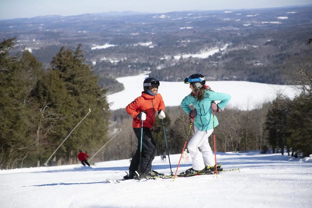 Male skier in orange ski jacket and black ski pants and female skier in light blue ski jacket and white ski pants hitting the slopes at Wachusett Mountain in Massachusetts, USA.