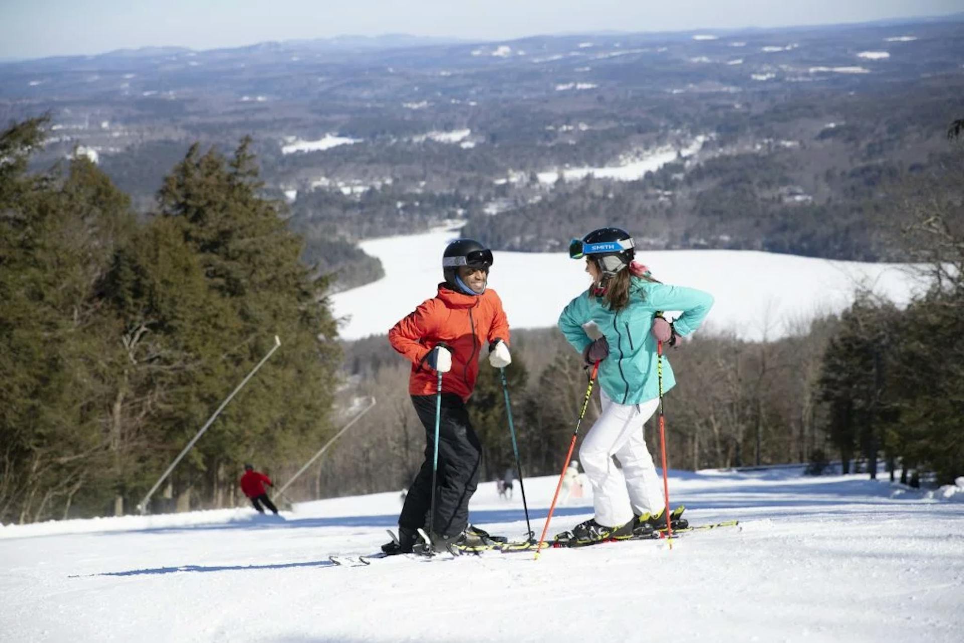 Male skier in orange ski jacket and black ski pants and female skier in light blue ski jacket and white ski pants hitting the slopes at Wachusett Mountain in Massachusetts, USA.