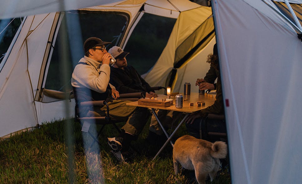 a photo of a group of people socializing at ta table in a shelter. one person is lifting a drink to his mouth.