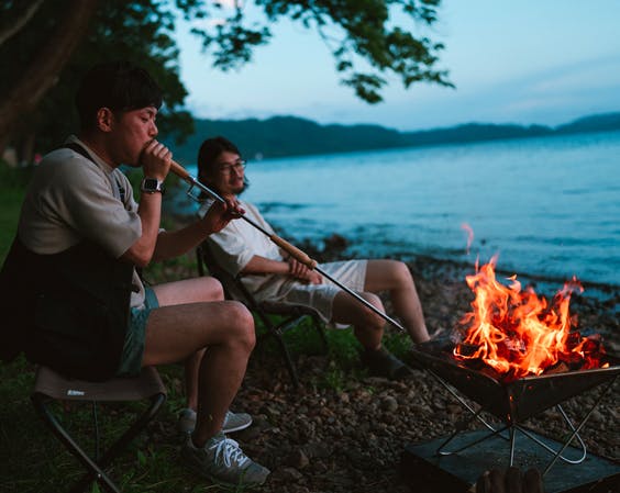 a photo of two people at dusk around a fire by the water and one is using a fire blower to stoke the flame