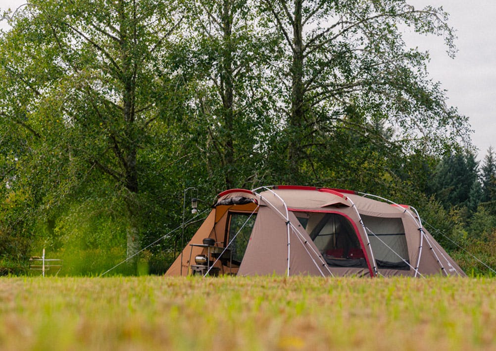 a photo of a brown Snow Peak Land station shelter in a field with trees behind it