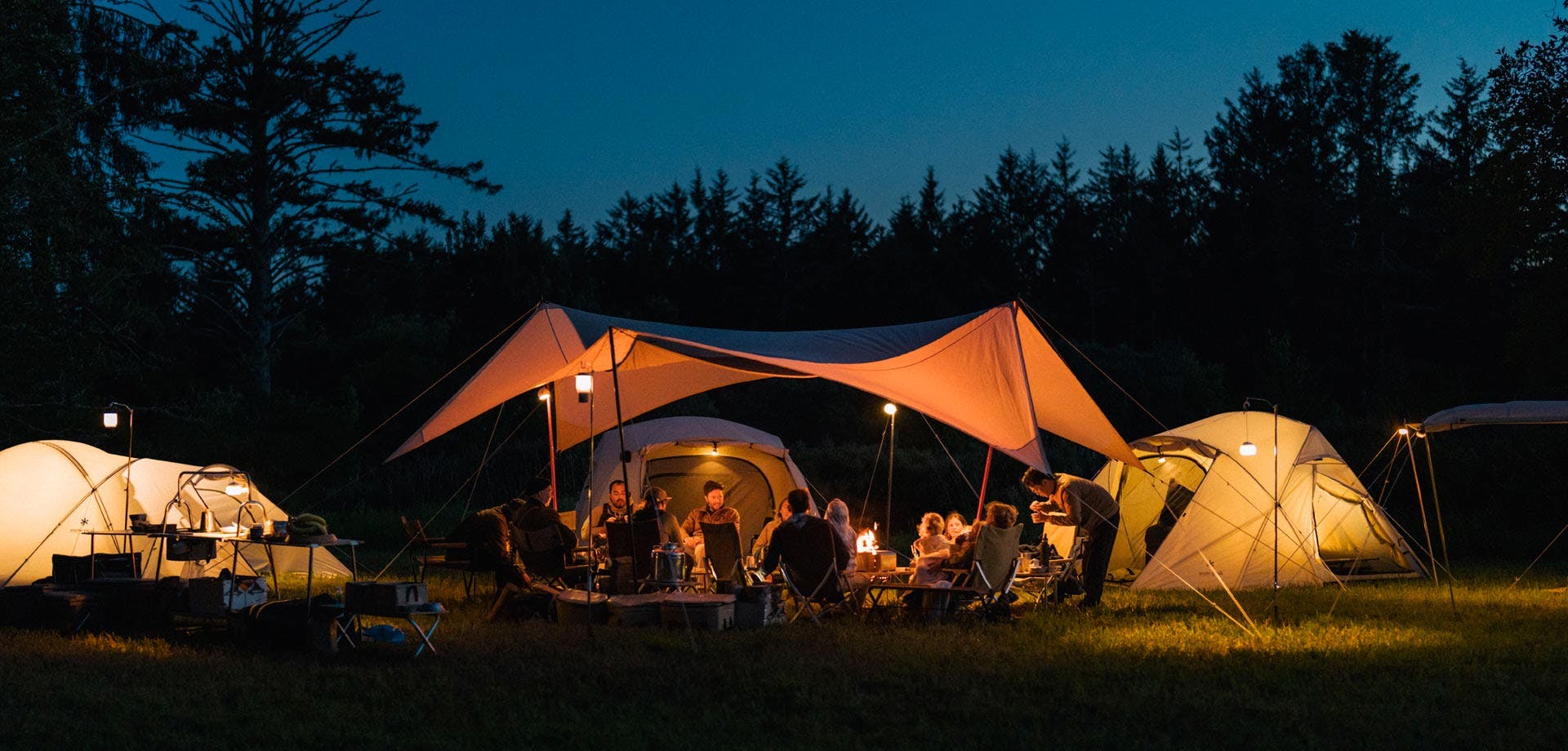 A photo of a group of 15-20 people around a campfire at night. They are under a tarp with ivory tents lit up around them.