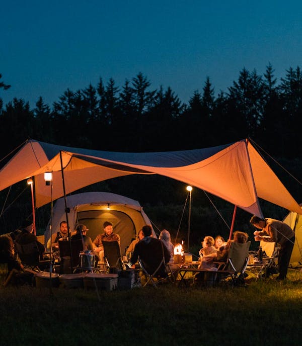 A photo of a group of 15-20 people around a campfire at night. They are under a tarp with ivory tents lit up around them.