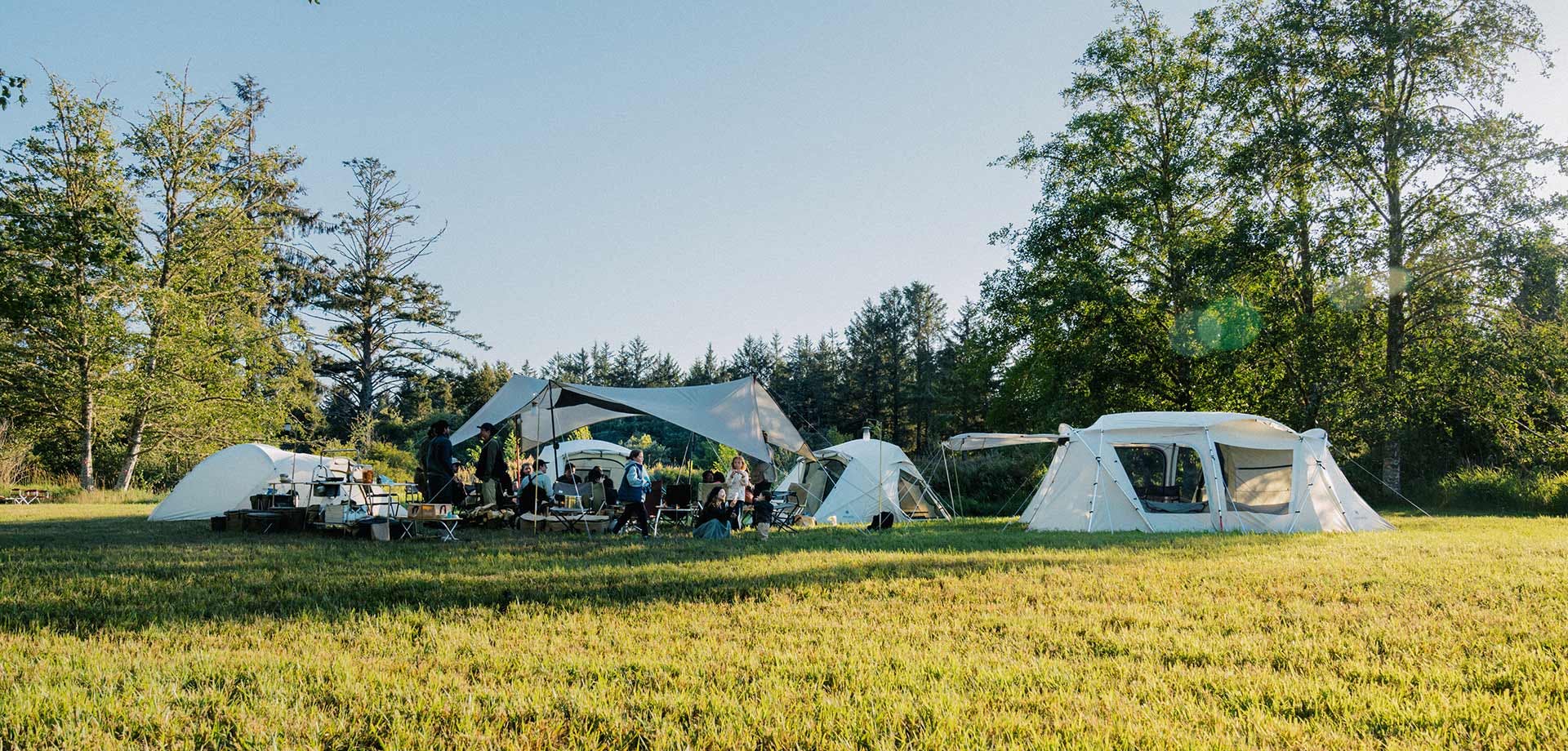 A full Snow Peak style campsite with four ivory tents surrounding a large ivory tarp with campers gathered beneath.