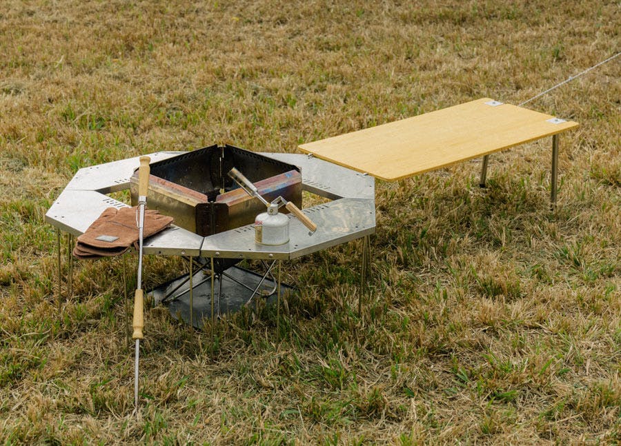 A photo of a snow Peak fire pit with a metal surrounding table and wooden extension table. It has gloves and fire tools with it.