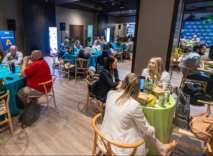 The image shows a conference breakout session with attendees seated at round tables covered in teal tablecloths. Participants appear to be engaged in discussions or networking, with some individuals facing each other in conversation, while others sit alone, possibly taking a break or awaiting the next session. In the background, promotional banners with logos are visible, indicating the event might have multiple sponsors or exhibitors. The setting has an informal and collaborative atmosphere, conducive to networking and small group interactions.