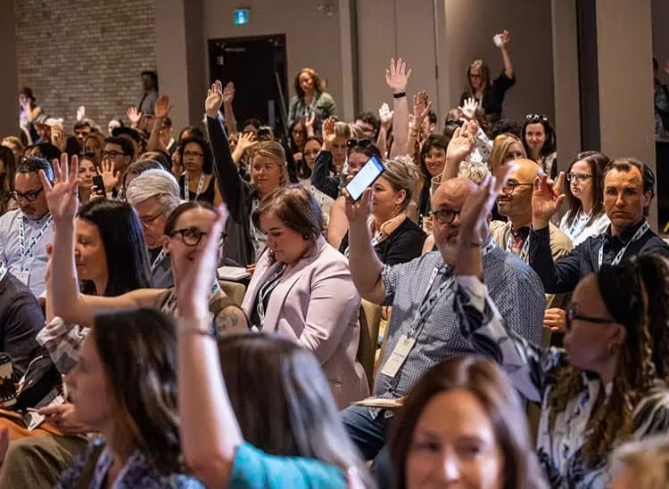 The image captures a conference room filled with attendees, many of whom have their hands raised, indicating participation in a Q&A session, a vote, or a show of agreement with the speaker. The audience appears attentive and engaged, with some individuals looking towards the front of the room where the speaker might be located. The atmosphere seems interactive and dynamic, with a clear focus on the event proceedings.