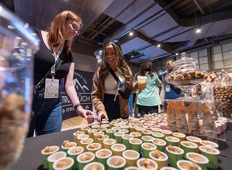 The image shows two individuals at a sampling station, one selecting a sample. In the foreground, multiple small cups with tastings are neatly arranged on a table. There's a display of snacks in glass jars, indicating a variety of offerings. The background features a banner with the text 