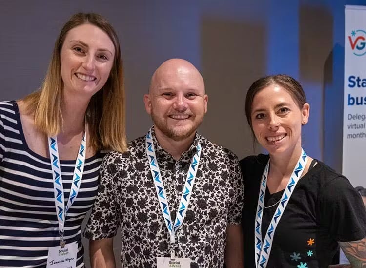  The image features three individuals smiling for the camera, two women and one man, all wearing lanyards with badges that indicate they might be at a social or professional event. The badges suggest they are either attendees or organizers of the event. They are dressed in business casual attire; the man is wearing a patterned shirt, and the women are in solid-colored tops. A banner in the background hints at a corporate or conference setting, creating a professional yet friendly atmosphere.