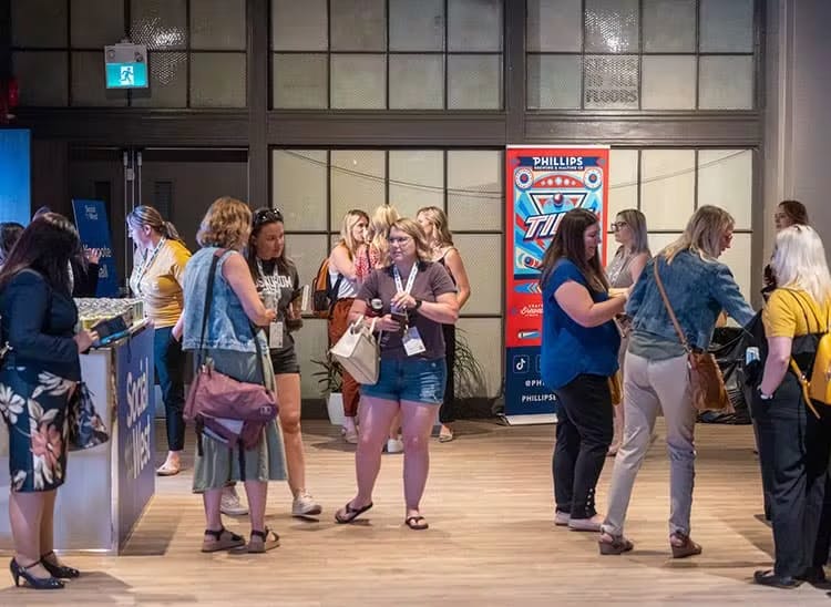 Attendees engaging at the SocialWest event registration area. A group of professionals are gathered in conversation, some are registering at the front desk staffed by a team member. The venue features industrial-style windows and a branded vending machine in the background, creating an informal yet energetic networking atmosphere