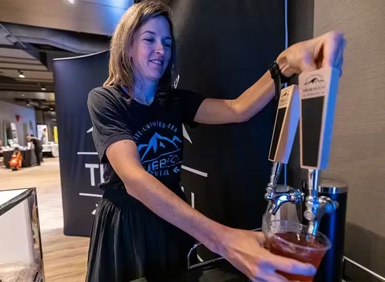 The image shows a woman smiling as she pours a drink from a tap, which appears to be housed in a large, dark-colored dispenser. She is wearing a casual black T-shirt with a graphic of mountains and the text