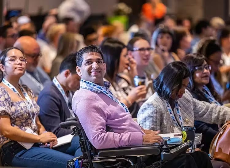 The image shows a diverse group of attendees at an event, focusing on a man in a wheelchair in the foreground. He is smiling and looking up, possibly at a speaker or a presentation, indicating engagement and interest. The audience around him is similarly attentive, with some people taking notes and others looking towards the stage. The environment suggests an inclusive and accessible conference setting, where all participants are involved in the learning experience
