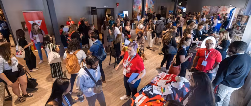 The image shows a bustling conference or event hall where attendees are networking. The crowd is diverse, with people wearing conference badges, indicating that they might be participants, speakers, or exhibitors. Some individuals are standing at booths, suggesting that the event includes vendor or information stations. There's an active exchange of ideas and information taking place, with some attendees carrying promotional materials. This environment is typical of industry conferences, trade shows, or professional gatherings, where participants engage in learning, networking, and promotional activities.
