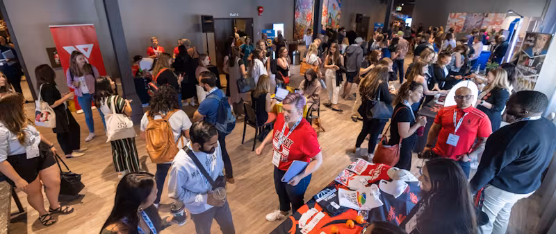 The image shows a bustling conference or event hall where attendees are networking. The crowd is diverse, with people wearing conference badges, indicating that they might be participants, speakers, or exhibitors. Some individuals are standing at booths, suggesting that the event includes vendor or information stations. There's an active exchange of ideas and information taking place, with some attendees carrying promotional materials. This environment is typical of industry conferences, trade shows, or professional gatherings, where participants engage in learning, networking, and promotional activities.