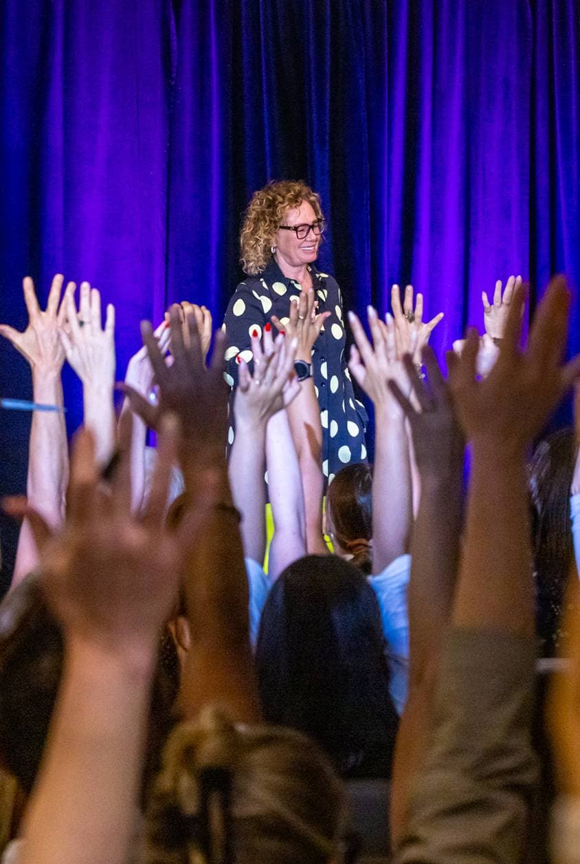 In the image, a speaker stands on a stage with a microphone in hand, addressing an audience. The attendees appear to be actively engaged, with several hands raised in the air, possibly in response to a question from the speaker or to volunteer for an interactive segment of the presentation. The speaker is smiling and seems to be interacting with the audience members. The backdrop of the stage is a simple, solid-colored curtain, focusing the audience's attention on the speaker. This setting is typical of a conference or seminar where there is direct interaction between the presenter and the audience.