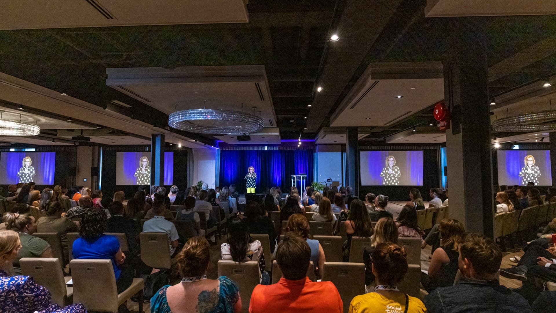  The image captures an indoor conference scene with an audience seated in rows, attentively facing a speaker on stage. The speaker, visible on two large screens for better visibility, stands at a lectern against a backdrop of blue curtains illuminated by stage lights. The room is well-lit, with elegant chandeliers overhead, and the audience appears engaged in the presentation, indicative of a professional or educational event.