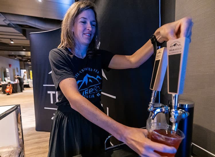  The image shows a woman smiling as she pours a drink from a tap, which appears to be housed in a large, dark-colored dispenser. She is wearing a casual black T-shirt with a graphic of mountains and the text 